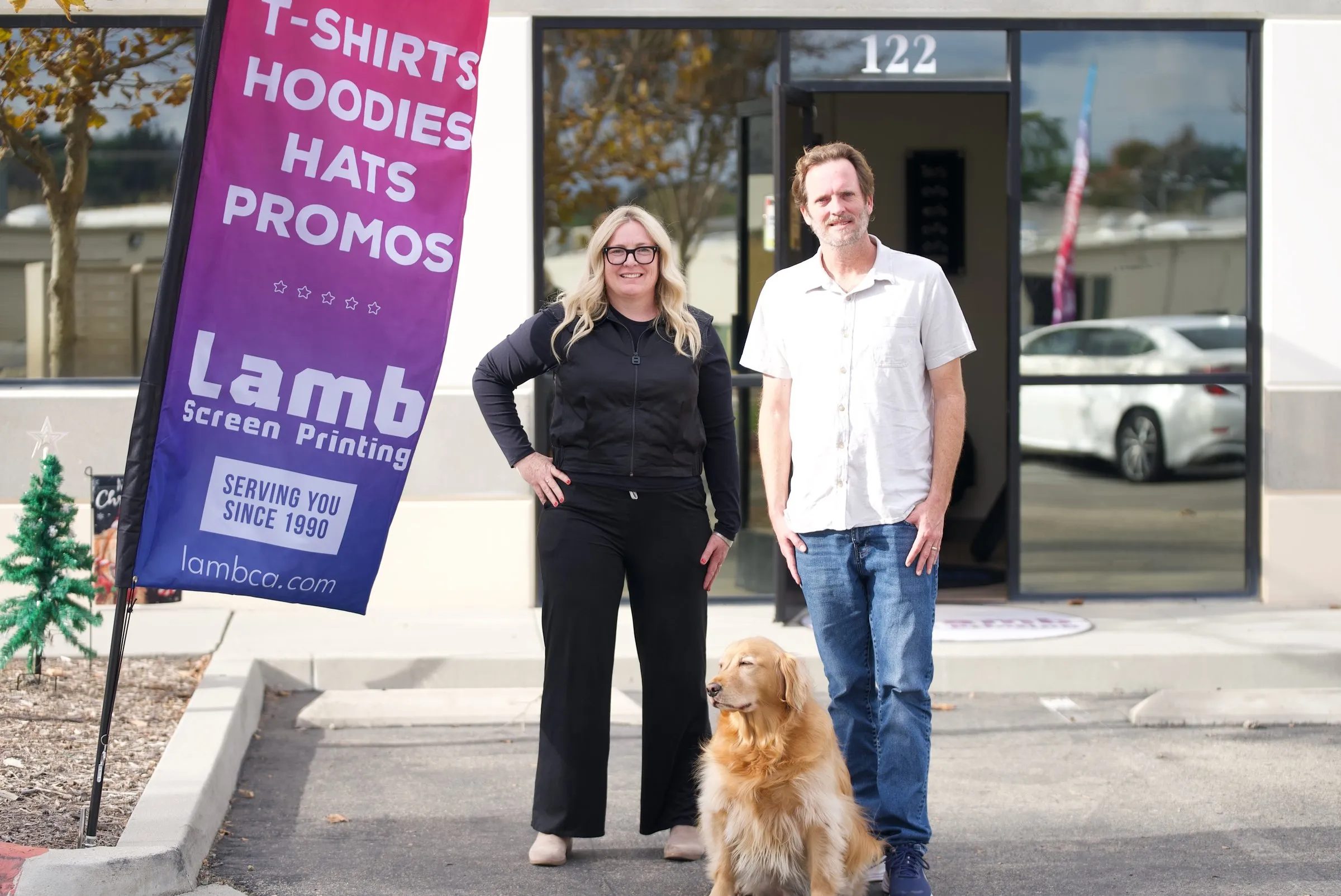 Two owners and their dog outside Suite 122 of the shop, beside the shop's flag for T-shirts, hoodies, hats and promos