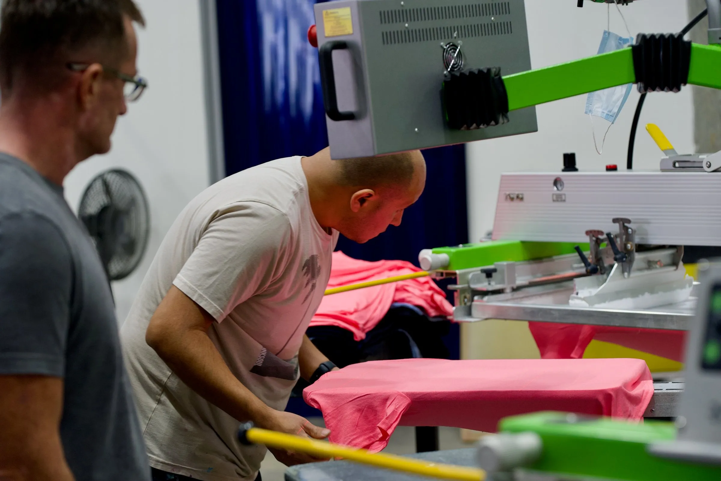 A printer lines up a pink shirt on a screen printing press while a coworker watches