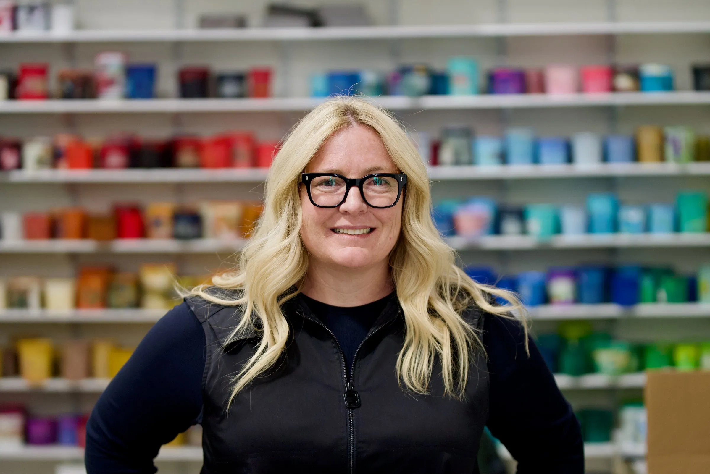 A member of the Lamb Screen Printing team in glasses and a black vest, in front of the shop's ink wall