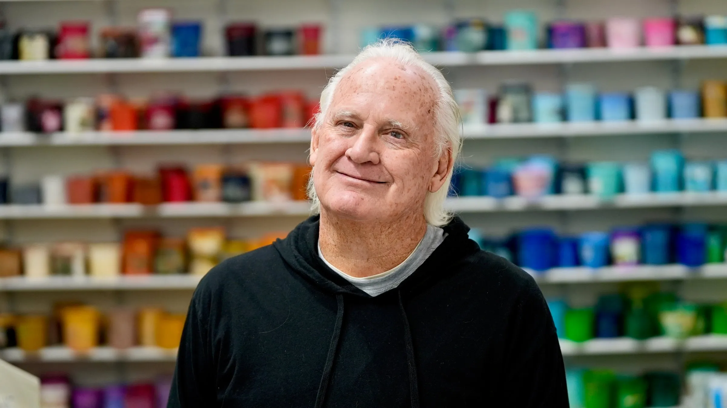 A member of the Lamb Screen Printing team with white hair and a black hoodie, in front of the shop's ink wall