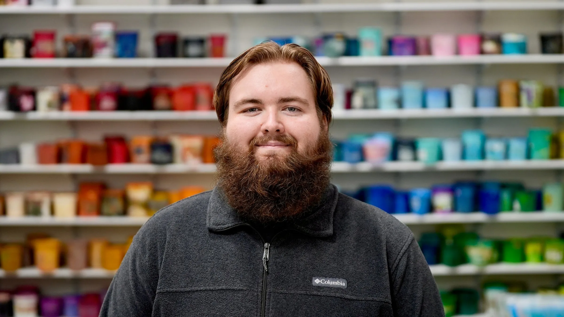 A member of the Lamb Screen Printing team with a red beard and a gray fleece, in front of the shop's ink wall