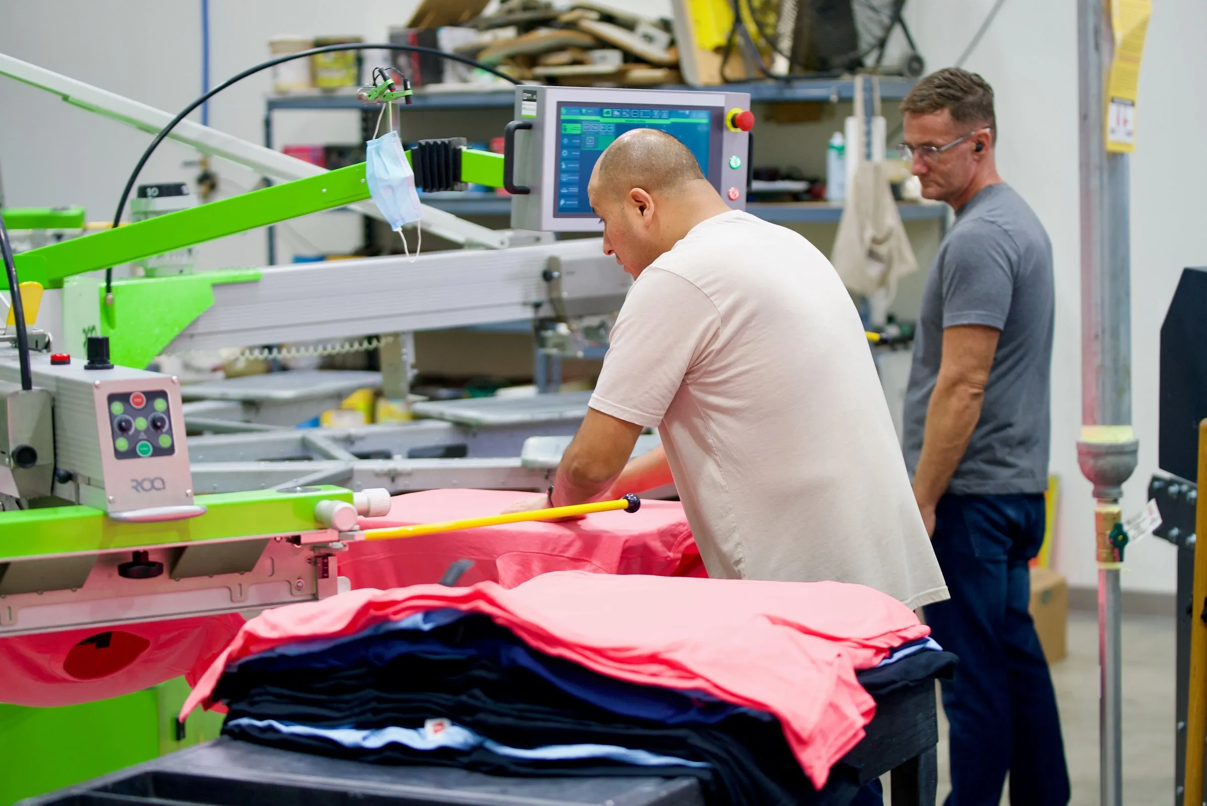 A printer smooths a pink shirt onto a platen of the automatic press while a coworker looks on