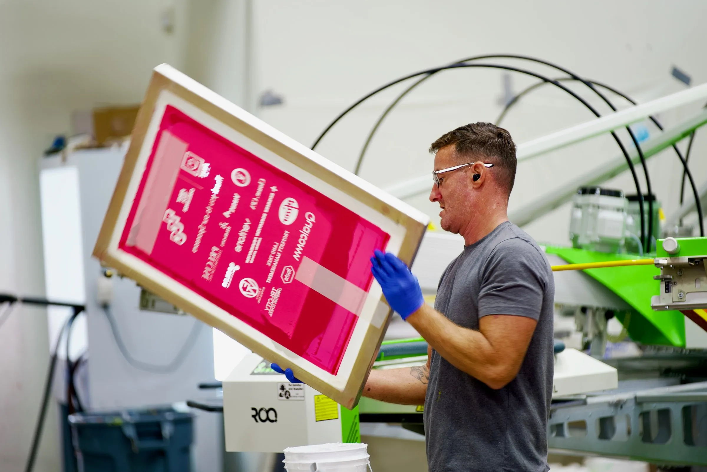 A printer in safety glasses and blue gloves holds up a freshly coated pink screen beside the automatic press