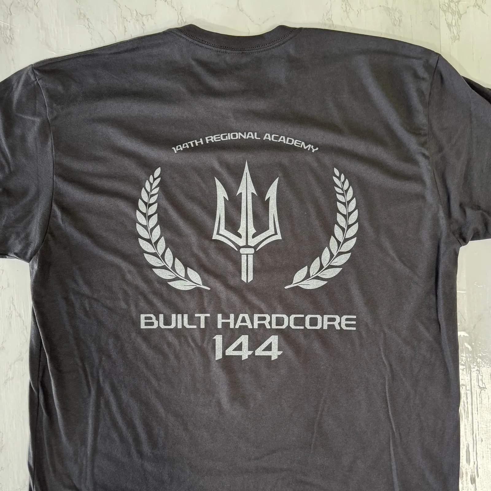 Back of a charcoal tee with a light gray trident and laurel print