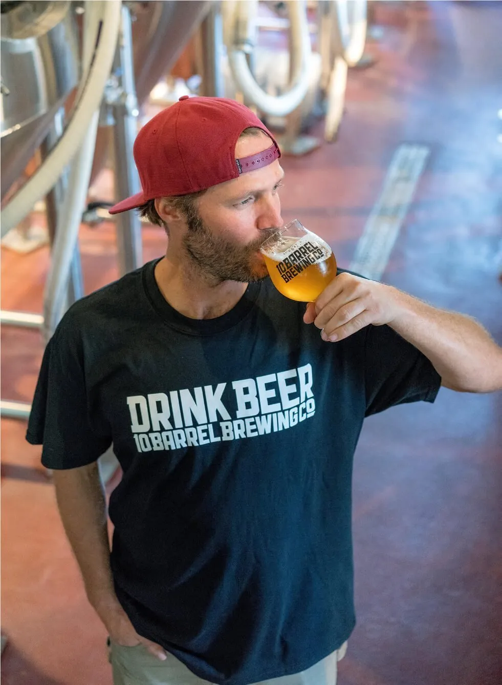 A man wearing a "drink beer 10 barrel brewing" t-shirt is tasting beer in a brewery.