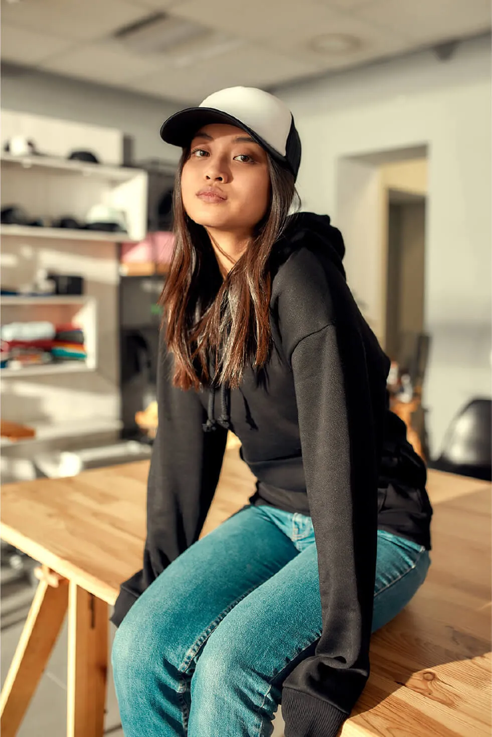 Woman sitting on a wooden table wearing a baseball cap and hoodie.
