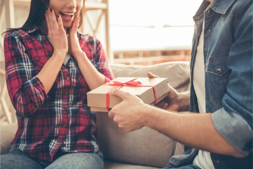 A person presenting a gift box with a red ribbon to a delighted woman.