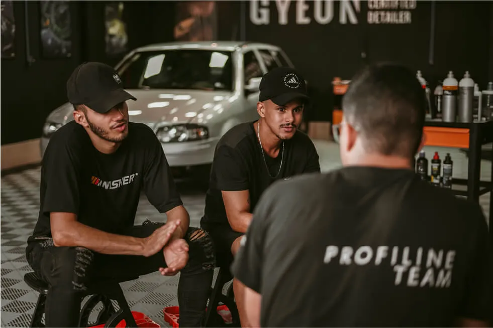 Three individuals in a workshop engaged in a conversation, with car detailing products on the shelf in the background.
