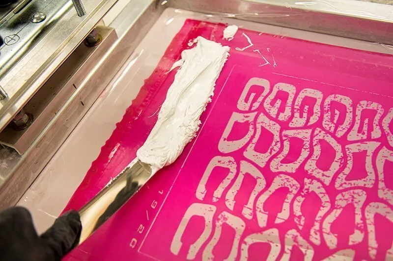 Screen printing process with squeegee and white ink on a pink stencil.