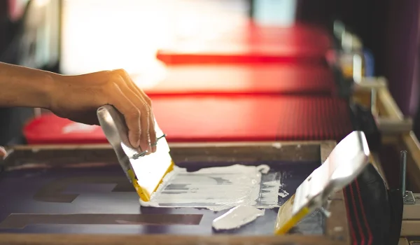 A person's hand using a squeegee to spread ink on a screen printing frame.