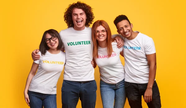 Four happy volunteers wearing white t-shirts with the word "volunteer" standing together against a yellow background.