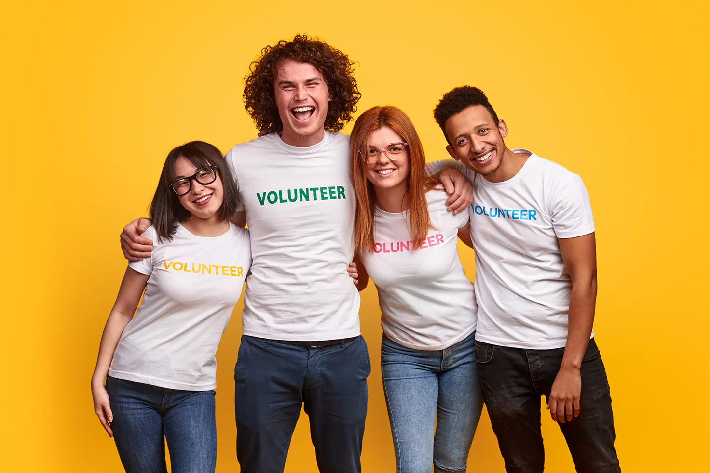 Four happy volunteers wearing white t-shirts with the word "volunteer" standing together against a yellow background.